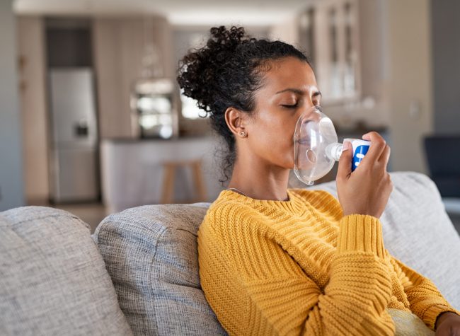 Black young woman holding a mask nebulizer inhaling fumes medication into lungs. African sick lady inhaling through inhaler mask sitting on the couch with copy space. Self treatment of the respiratory tract using inhalation nebulizer.