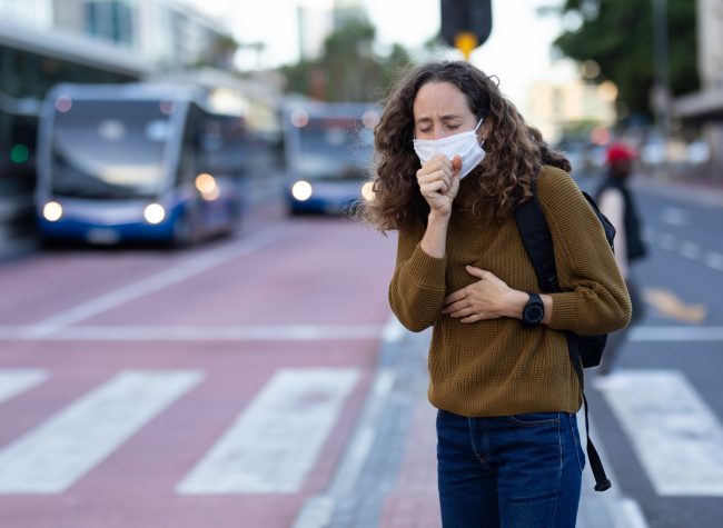 Caucasian woman out and about in the city streets during the day, wearing a face mask against covid19 coronavirus covering her face while coughing
